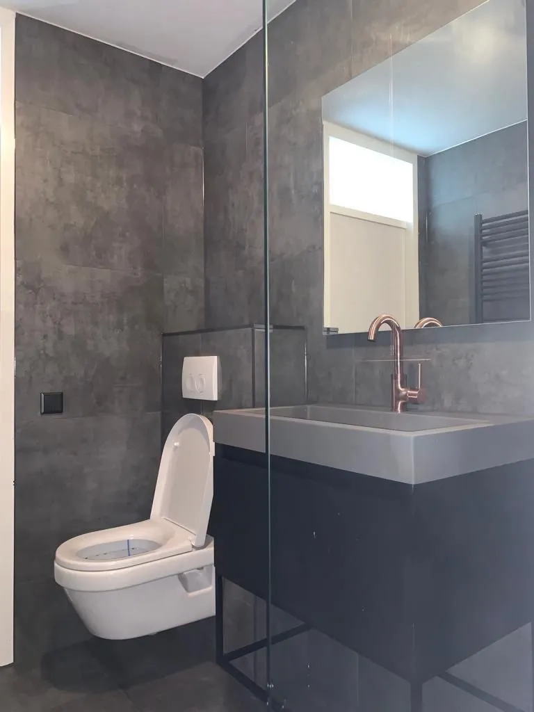 Badkamer met grijze tegels, hangtoilet en wastafel op een zwart onderstel (projectfoto bij review van Partoredjo)