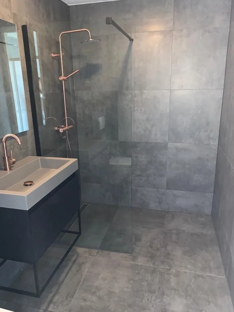 Grijs betegelde badkamer met inloopdouche, glaswand en wastafel met koperkleurige kraan (projectfoto bij review van Partoredjo)