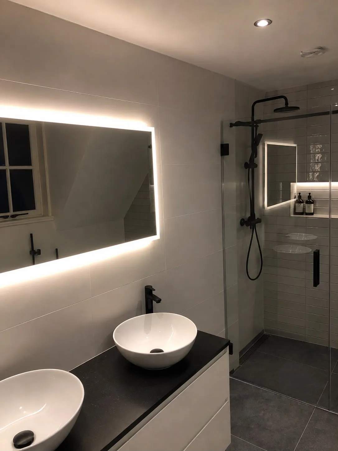 Badkamer met verlichte spiegel, witte waskom op zwart werkblad en een douche met zwarte kranen