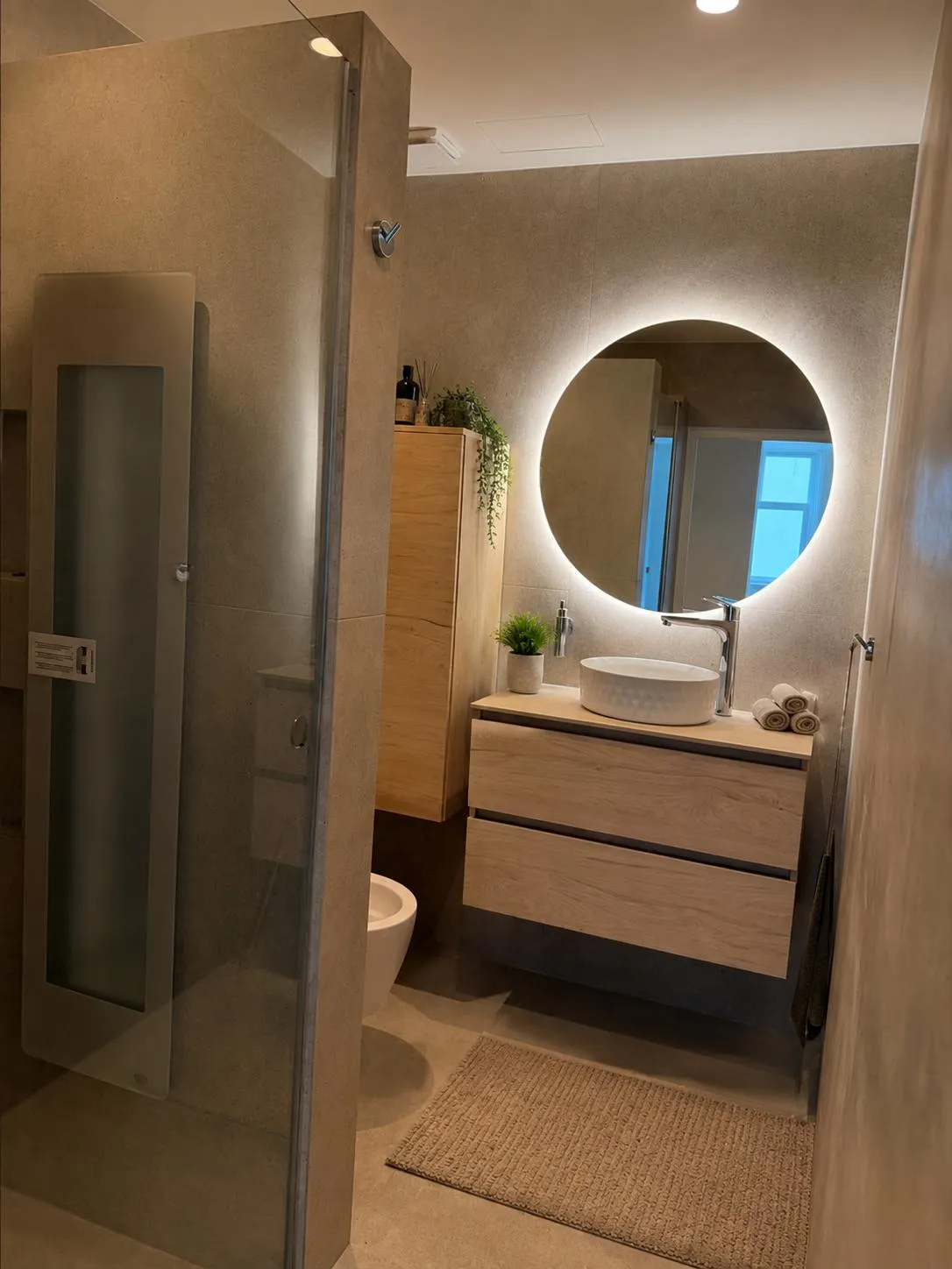 Badkamer met houten wastafelmeubel, ronde verlichte spiegel en glazen douchewand